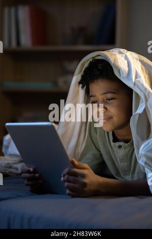 Content African American boy lying with tablet under duvet in dark nursery room Stock Photo