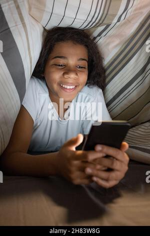 Smiling pretty African American girl hiding under duvet and using smartphone while texting online at night Stock Photo