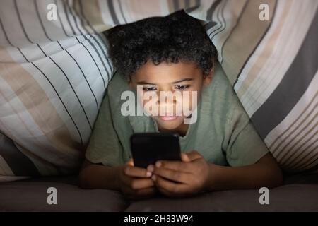 Content cute Black boy with curly hair lying on bed and using smartphone under duvet at night Stock Photo