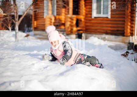 Little girl is lying on snowdrift. Child having fun on sunny winter day in backyard on background of wooden house, playing snowballs, jumping in snow Stock Photo
