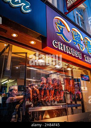 CRISPY DUCK WINDOW DISPLAY CHINATOWN SOHO LONDON Chef arranging rows of enticing Peking crispy duck and pork belly ribs hanging air drying in Chinese restaurant window display at dusk, Gerrard Street Chinatown Soho London Stock Photo