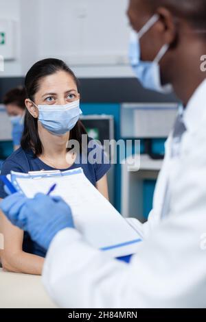 Closeup of african american pediatrician doctor writing ill diagnostic on clipboard explaining healthcare treatment to parent during clinical appointment in hospital office. Therapist with face mask Stock Photo