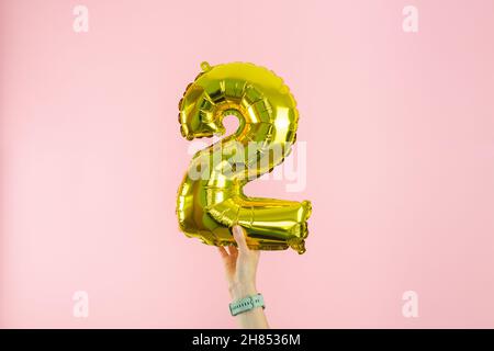 Woman's hand with watch holding golden globe in the shape of number 2 on pink background. Copy space. Stock Photo