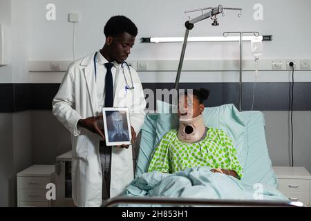 African american radiologist man holding tablet discussing bones radiography with sick woman during medical examination in hospital ward. Patient wearing neck cervical collar recovering after surgery Stock Photo