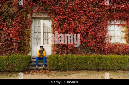 Young Spanish man sitting on the doorsteps and using a tablet; a house wall covered with ivy leaves Stock Photo
