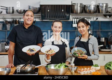 Waist up portrait of three women wearing aprons holding gourmet dishes in professional kitchen and smiling at camera Stock Photo