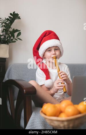 Boy in Santa hat learning to play the flute watching online course on laptop while practicing at home. Online training, online classes. Stock Photo