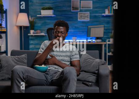 African american guy watching entertainment movie on television laughing during comedy film relaxing in living room. Black young man sitting on sofa while eating popcorn enjoying free time Stock Photo