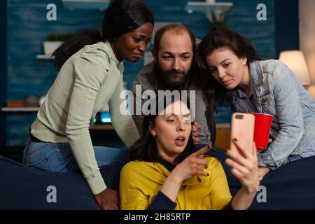 Multi-ethnic friends looking at fuuny videos on internet using smartphone during relaxing night at home enjoying hanging out together. Group of people having fun celebrating weekend in living room Stock Photo