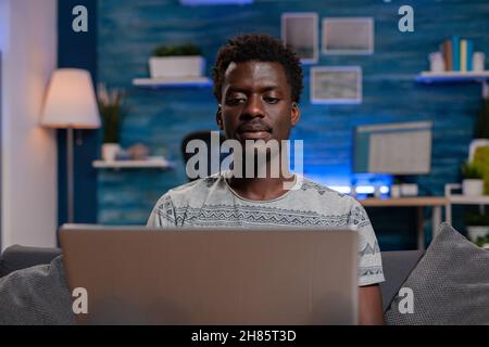 African american young businessman sitting on sofa browsing communication information working remote from home at virtual project. Black entrepreneur analyzing financial graph on laptop Stock Photo