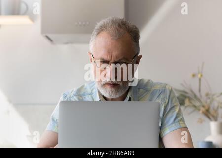 Focused older 60s man wearing glasses, working at laptop Stock Photo