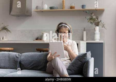 Cheerful elderly 60s woman in wireless headphones watching movie Stock Photo