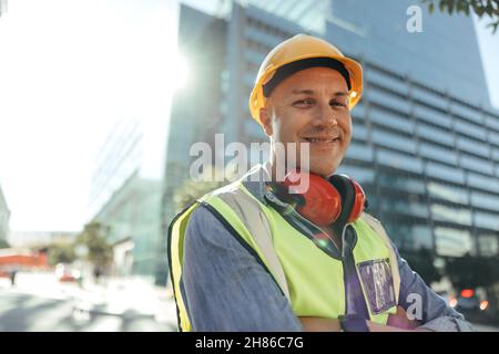 Happy construction worker smiling at the camera while standing with his arms crossed in the city. Mid-adult blue collar worker standing in front of hi Stock Photo