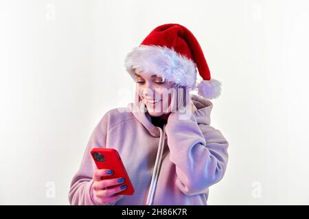 Amazed attractive young woman in santa claus hat with pink hair in lilac hoodie using cell phone over white background Stock Photo