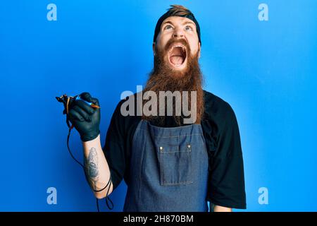Redhead man with long beard tattoo artist wearing professional uniform and gloves angry and mad screaming frustrated and furious, shouting with anger. Stock Photo
