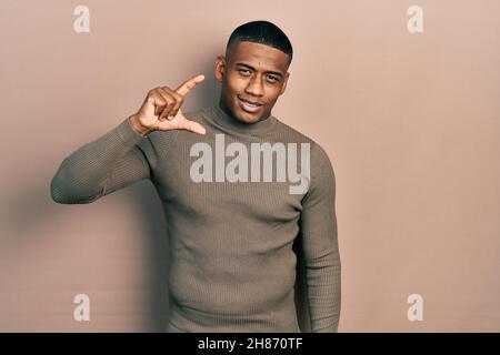 Young black man wearing casual turtleneck sweater smiling and confident gesturing with hand doing small size sign with fingers looking and the camera. Stock Photo