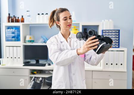 Young woman wearing scientist uniform holding virtual reality goggles at laboratory Stock Photo