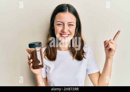 Young brunette woman holding soluble coffee smiling happy pointing with hand and finger to the side Stock Photo