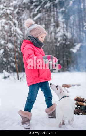 Little girl in a bright jacket plays in the winter snowy forest with her dog jack russell terrier Stock Photo