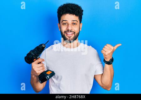 Young arab man with beard using drill pointing thumb up to the side smiling happy with open mouth Stock Photo