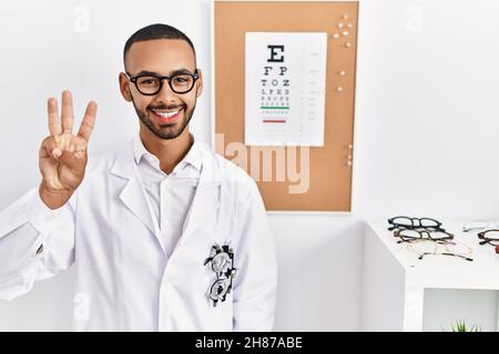 African american optician man standing by eyesight test showing and pointing up with fingers number three while smiling confident and happy. Stock Photo