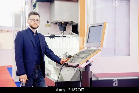 Engineer young man in glasses operates CNC automatic metalworking lathe or laser, industrial worker in modern factory. Stock Photo