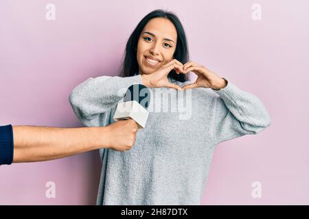 Young hispanic girl being interviewed by reporter holding microphone smiling in love doing heart symbol shape with hands. romantic concept. Stock Photo