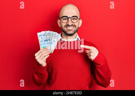 Young bald man holding peruvian sol banknotes smiling happy pointing with hand and finger Stock Photo