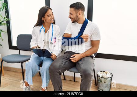 Youg latin injuried man wearing sling talking with the doctor at clinic waiting room. Stock Photo