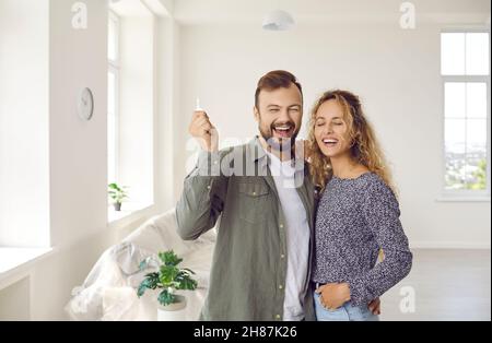 Happy young couple standing in their new house, showing keys to new home and laughing Stock Photo