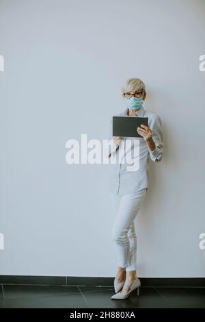 Mature woman with protective facial mask using digital tablet in the office Stock Photo