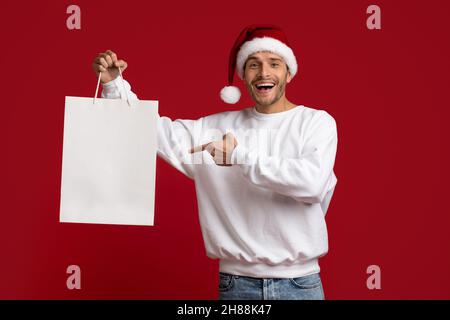 Shopping Offer. Excited Man In Santa Hat Pointing At Blank Shopper Bag Stock Photo
