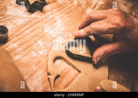 Making gingerbread cookie, cutting heart shape from dough by cookie cutter. Senior  woman making traditional gingerbread biscuit. Close-up old hand Stock Photo