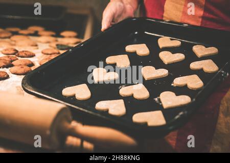 Gingerbread heart cookies on tray are ready for baking in oven. Woman making traditional Christmas cookies. Domestic kitchen. Toned image Stock Photo