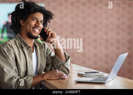 Black Freelancer Guy Talking On Cellphone And Working With Laptop At Cafe Stock Photo