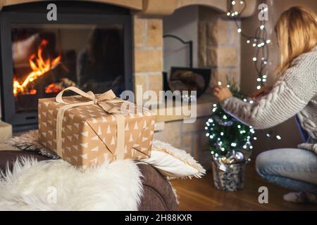 Woman preparing small christmas tree and lights for Christmas Holiday. Selective focus on gift box. Cozy living room with fireplace Stock Photo