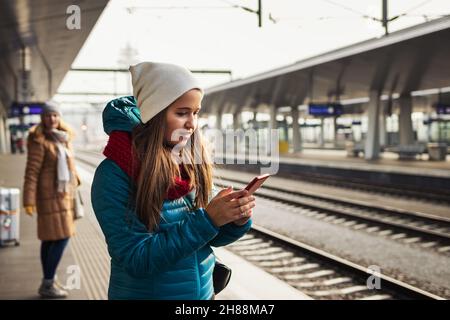 Woman is standing at railroad station and buying a railway ticket on her smart phone. People waiting for train on platform. Tourist using mobile app Stock Photo