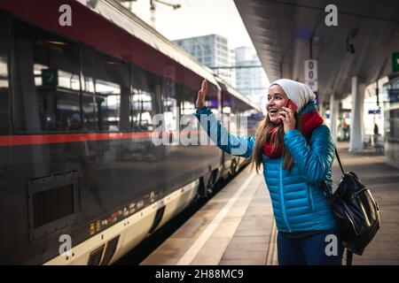 I can see you! Happy woman waving at passenger in the arriving train and talking to mobile phone. Smiling young woman looking to train at station Stock Photo
