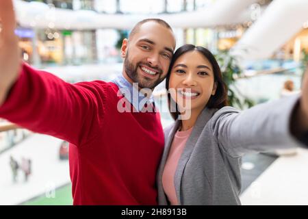 Loving young diverse couple smiling, taking selfie at huge shopping centre Stock Photo