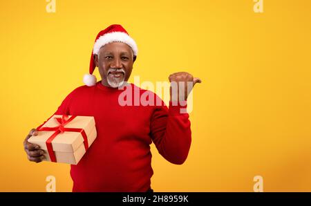 Cheerful senior black man holding Christmas gift, pointing aside at blank space on orange studio background Stock Photo