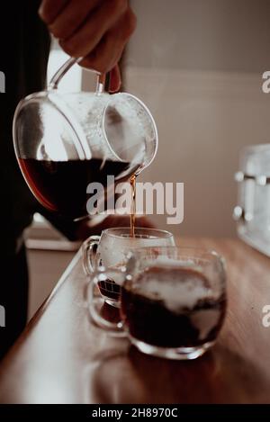 Young hipster mixed-race millennial man pouring freshly ground and brewed pour-over specialty coffee in his kitchen at home into glass novelty mugs Stock Photo