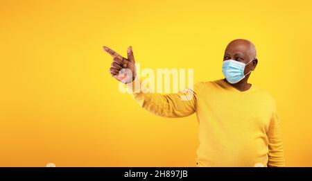 Elderly black man in surgical face mask pointing at blank space for text or ad over orange studio background, banner Stock Photo