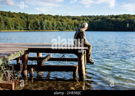 A man sits on the old wooden jetty by the lake in the afternoon and enjoys the peace and solitude. Stock Photo