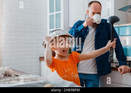 Happy daughter and father playing in kitchen in morning. Child kid girl put colander bowl on her head with ladle in hand she have fun and laughs Plays Stock Photo