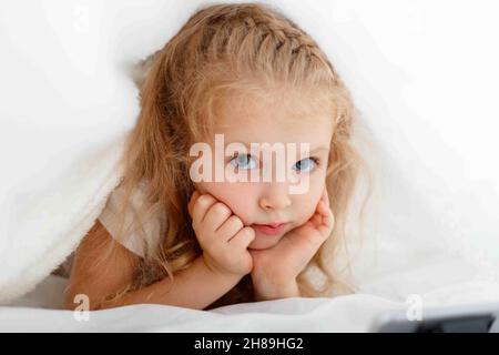 Portrait of beautiful caucasian 5 years girl child hiding in bed. Blonde Kid boring and hides under the cover to watch videos on smartphone at home.  Stock Photo