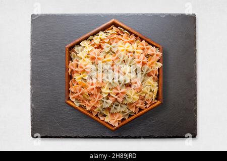 Slate tray on a white surface with on its middle, an hexagonal wooden dish with colorful bow tie pasta. Stock Photo