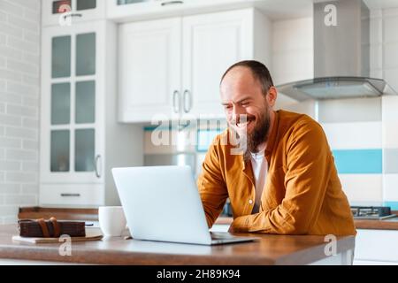 Young man working using laptop in kitchen in home interior. Smiling Freelancer works remotely from home. Happy man owner is doing paperwork using Stock Photo