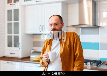 Portrait young happy man drinking coffee or tea in kitchen at home interior. Bald bearded 30s man enjoys morning routine in kitchen apartment.  Stock Photo