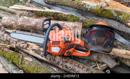 Portable power chain saw and safety helmet on heap of mossy wood logs. Orange chainsaw, hard hat, face guard and hearing protection. Sawing machine. Stock Photo