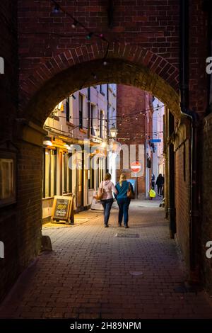 People walking at night through a brick archway on Church Street, a narrow alleyway that runs via College Street and King Street. Ludlow, England Stock Photo